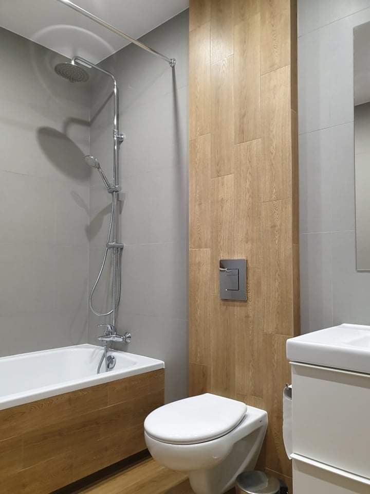 Nowoczesna łazienka z szarą glazurą i drewnopodobnymi akcentami: obudowa wanny i pionowy pas płytek imitujących drewno. Widoczna bateria prysznicowa, toaleta podwieszana i umywalka z szafką.
