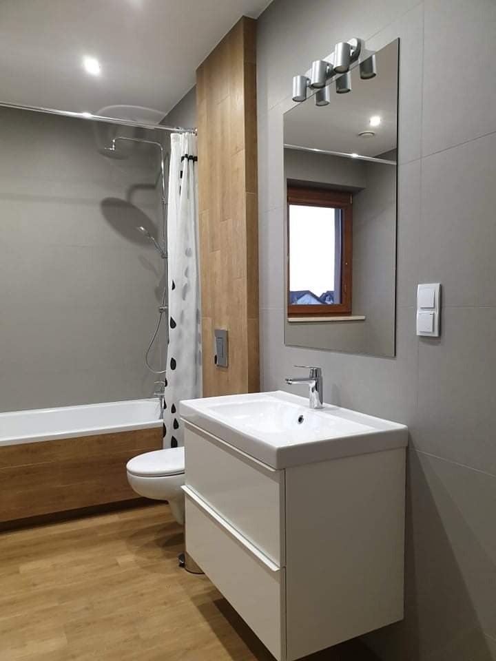 Nowoczesna łazienka z drewnianymi akcentami: obudowa wanny i panel ścienny, szare płytki, biała umywalka z szafką, lustro z oświetleniem punktowym, kabina prysznicowa z zasłoną w czarne krople.