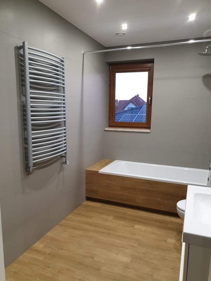 Jasna łazienka z drewnopodobną podłogą, grzejnikiem drabinkowym na ścianie, zabudowaną wanną z drewnianą obudową i oknem z widokiem na dom z panelami słonecznymi.