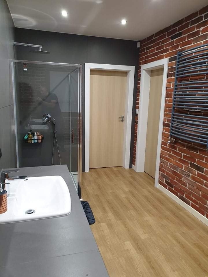 Nowoczesna łazienka z kabiną prysznicową typu walk-in, umywalką nablatową na szarym blacie, grzejnikiem drabinkowym na ścianie z czerwonej cegły oraz jasnymi panelami podłogowymi.