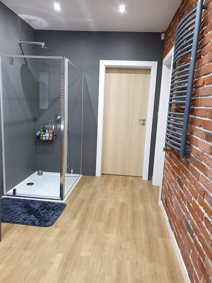 Nowoczesna łazienka z kabiną prysznicową typu walk-in, drewnopodobną podłogą, szarą ścianą i ścianą z czerwonej cegły z grzejnikiem drabinkowym.