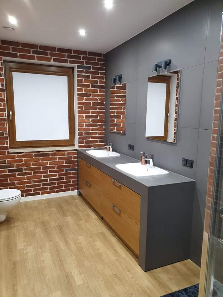 Nowoczesna łazienka z dwiema umywalkami nablatowymi na drewnianej szafce z szarym blatem, ceglana ściana z oknem i szara ściana z lustrami, drewniana podłoga, widoczna toaleta po lewej.