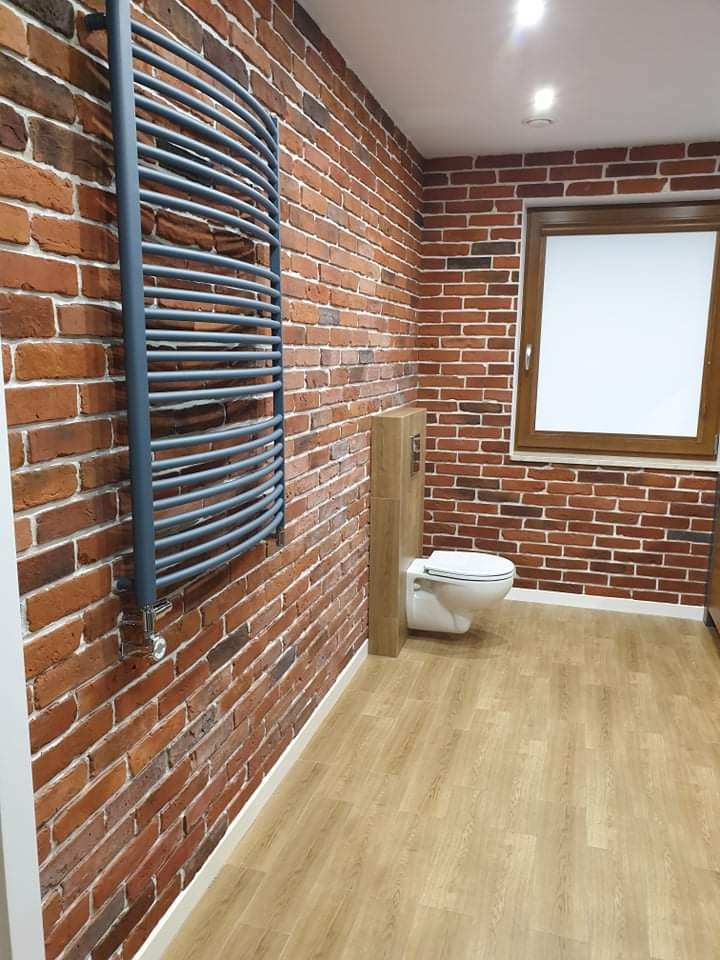 Łazienka z ceglaną ścianą, drewnianą podłogą i grzejnikiem drabinkowym w kolorze szarym, widoczne sanitariaty i okno z drewnianą ramą.