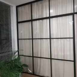 Białe, delikatne firany za czarną, metalową konstrukcją z podziałami, obok okno z żaluzjami i zielona roślina doniczkowa na parkiecie.