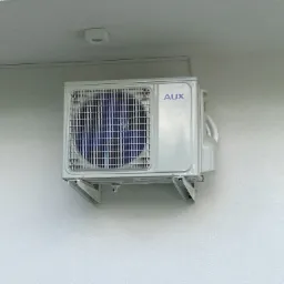 Zewnętrzna jednostka klimatyzacji marki AUX zamontowana na białej, teksturowanej ścianie budynku, widoczne elementy mocujące i fragment okna z żaluzjami.