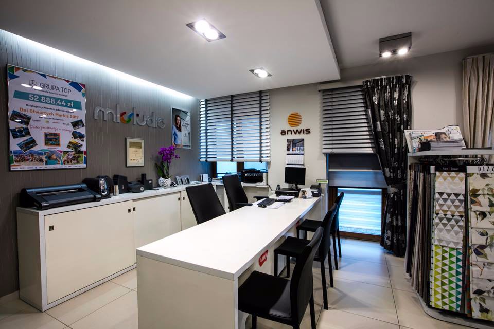 Wnętrze salonu firmowego z ekspozycją poziomych przesłon okiennych i wzorników tkanin zasłonowych, oświetlone lampami sufitowymi.