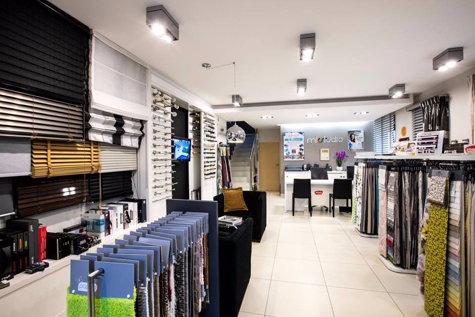 Wnętrze salonu z ekspozycją różnych typów osłon okiennych: drewniane, aluminiowe, rolety dzień i noc, oraz wzorniki tkanin na stojakach, biuro obsługi klienta w tle.