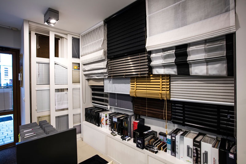 Ekspozycja różnorodnych osłon okiennych w salonie: poziome listwy w odcieniach brązu, szarości i czerni, rolety rzymskie z tkanin w paski i wzory, oraz katalogi z próbkami materiałów na półce.
