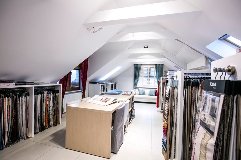 Salon z ekspozycją tkanin i próbek materiałów na rolety i zasłony, w jasnym wnętrzu na poddaszu ze skośnym sufitem i oknami dachowymi.