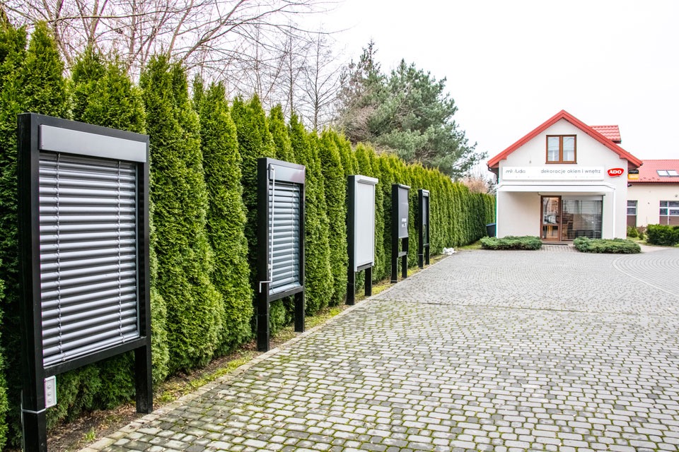 Ekspozycja zewnętrzna z różnymi modelami osłon okiennych na metalowych stojakach, ustawiona wzdłuż żywopłotu przed budynkiem firmy z szyldem 'Dekoracje okien i wnętrz'.