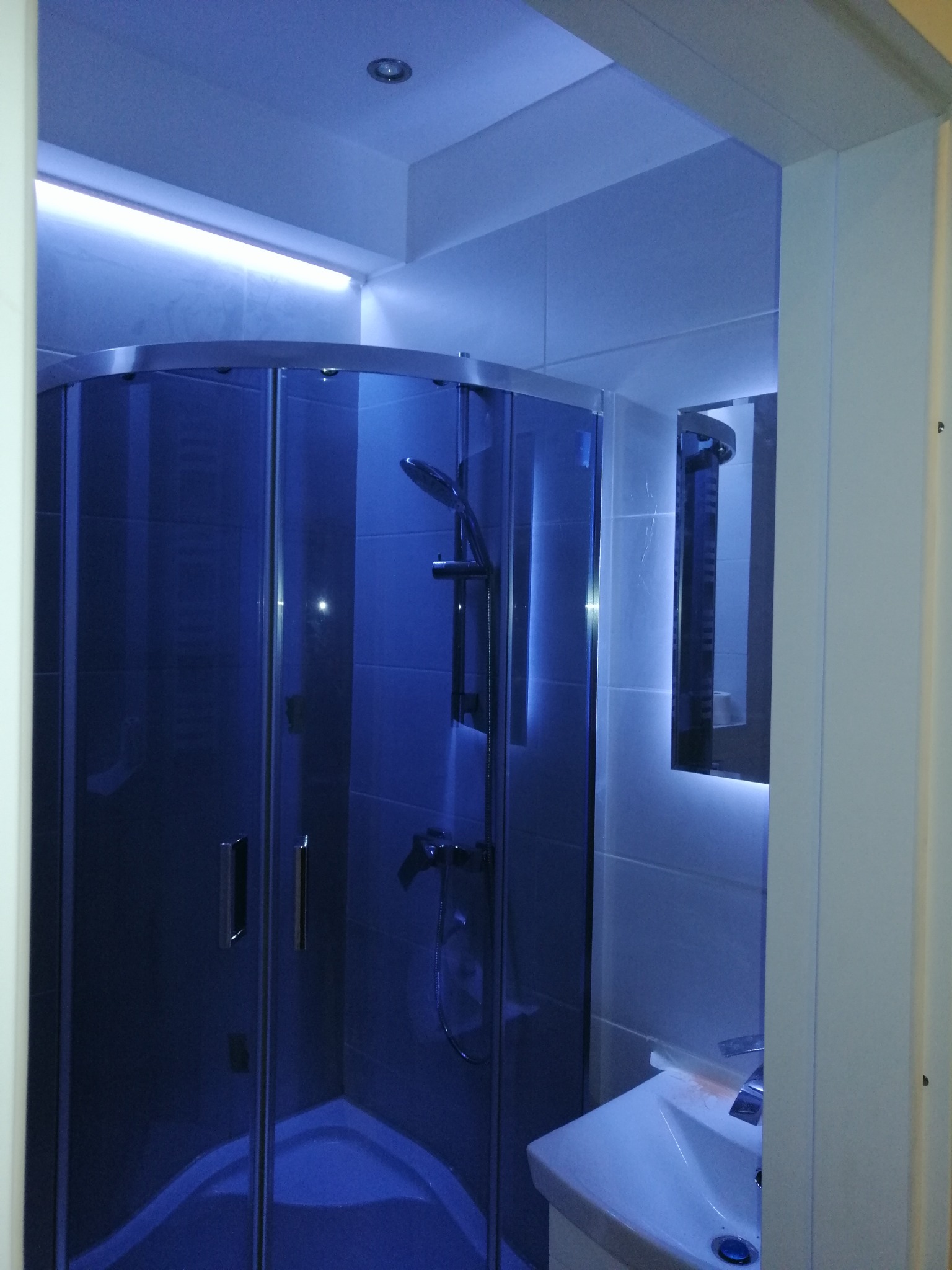 Nowoczesna kabina prysznicowa z hydromasażem, oświetlona niebieskim światłem LED, umywalka z baterią w minimalistycznej łazience z białymi płytkami.