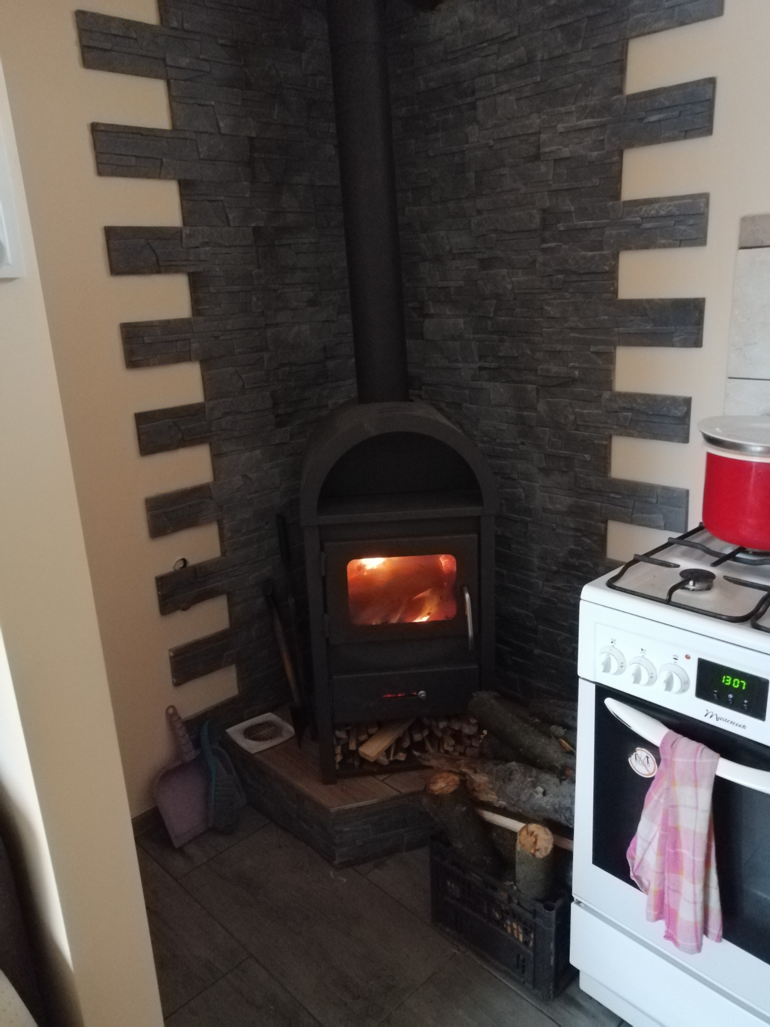 Czarne żeliwne palenisko z płomieniami, otoczone dekoracyjną ścianą z szarej cegły, obok biała kuchenka z czerwoną emaliowaną patelnią.