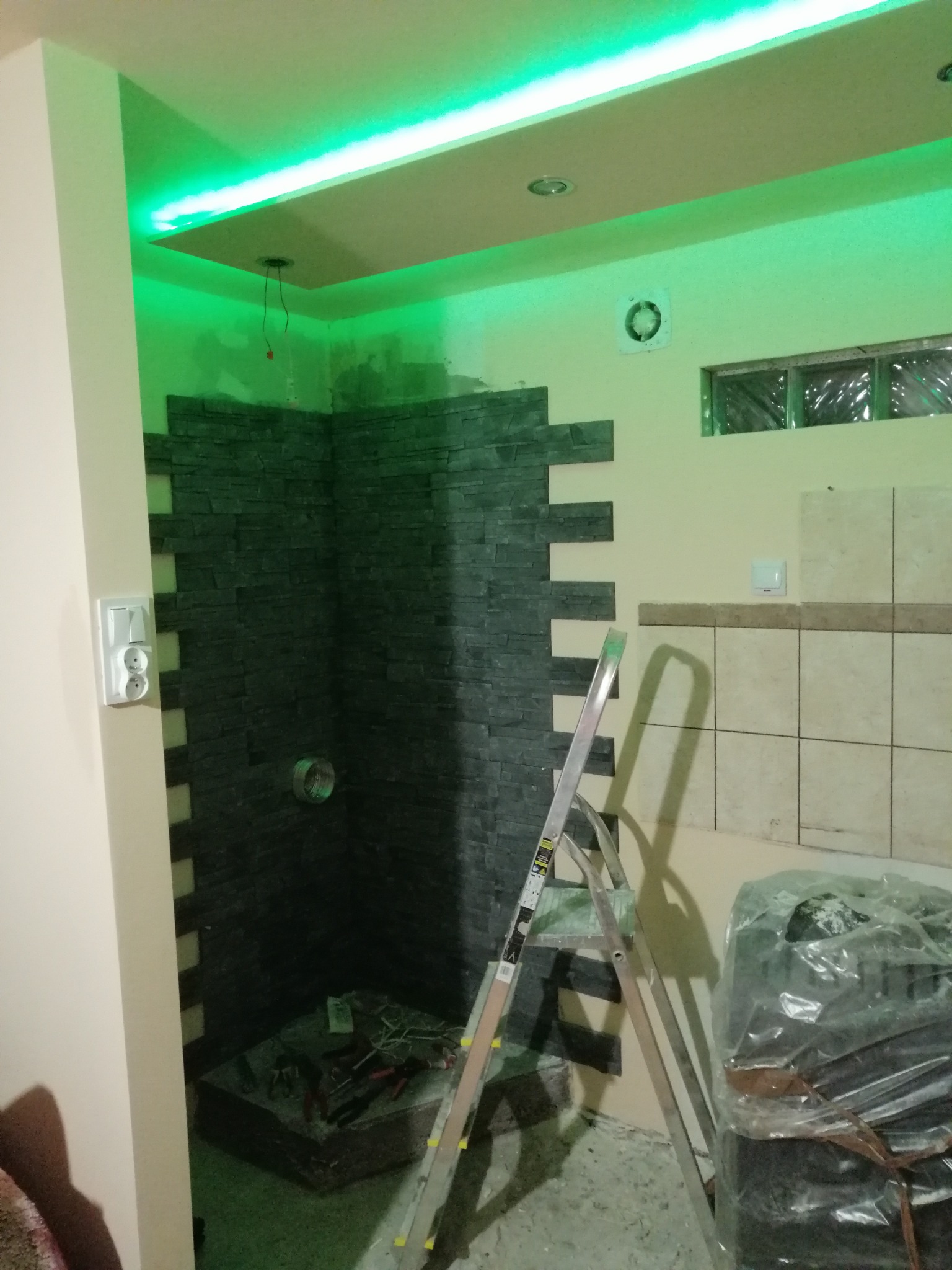Montaż dekoracyjnej, ciemnej okładziny kamiennej na ścianie w rogu łazienki, widoczna drabina i narzędzia, oświetlenie sufitowe w kolorze zielonym.