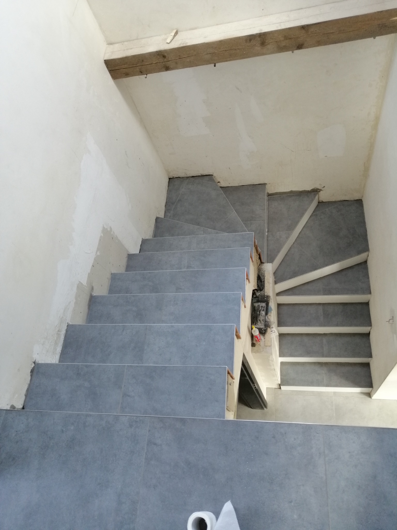 Wykończone płytkami schody w szarym odcieniu, widok z góry na nierówną ścianę i surowy strop drewniany.