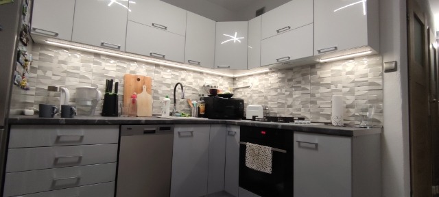 Nowoczesna kuchnia z białymi szafkami, szarym blatem i mozaiką na ścianie, oświetlenie LED pod szafkami górnymi, widok na kuchenkę, zlew i akcesoria kuchenne.