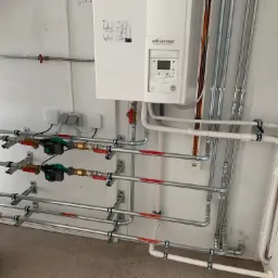 Węzeł cieplny z pompą ciepła, licznymi rurami i zaworami, zainstalowany w pomieszczeniu technicznym z białymi ścianami i szarą podłogą. Widoczny model pompy ciepła Waterstage.