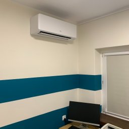 Klimatech Marcin Puc - Biały klimatyzator Fujitsu zamontowany na ścianie w biurze z poziomymi pasami w kolorze białym i turkusowym, widoczny komputer i drukarka na biurku.