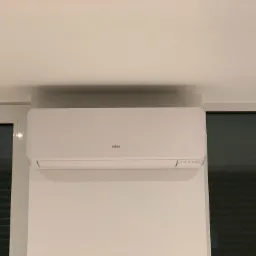 Biała jednostka wewnętrzna klimatyzacji marki Fujitsu zamontowana na ścianie w pomieszczeniu z oknem i drzwiami balkonowymi.