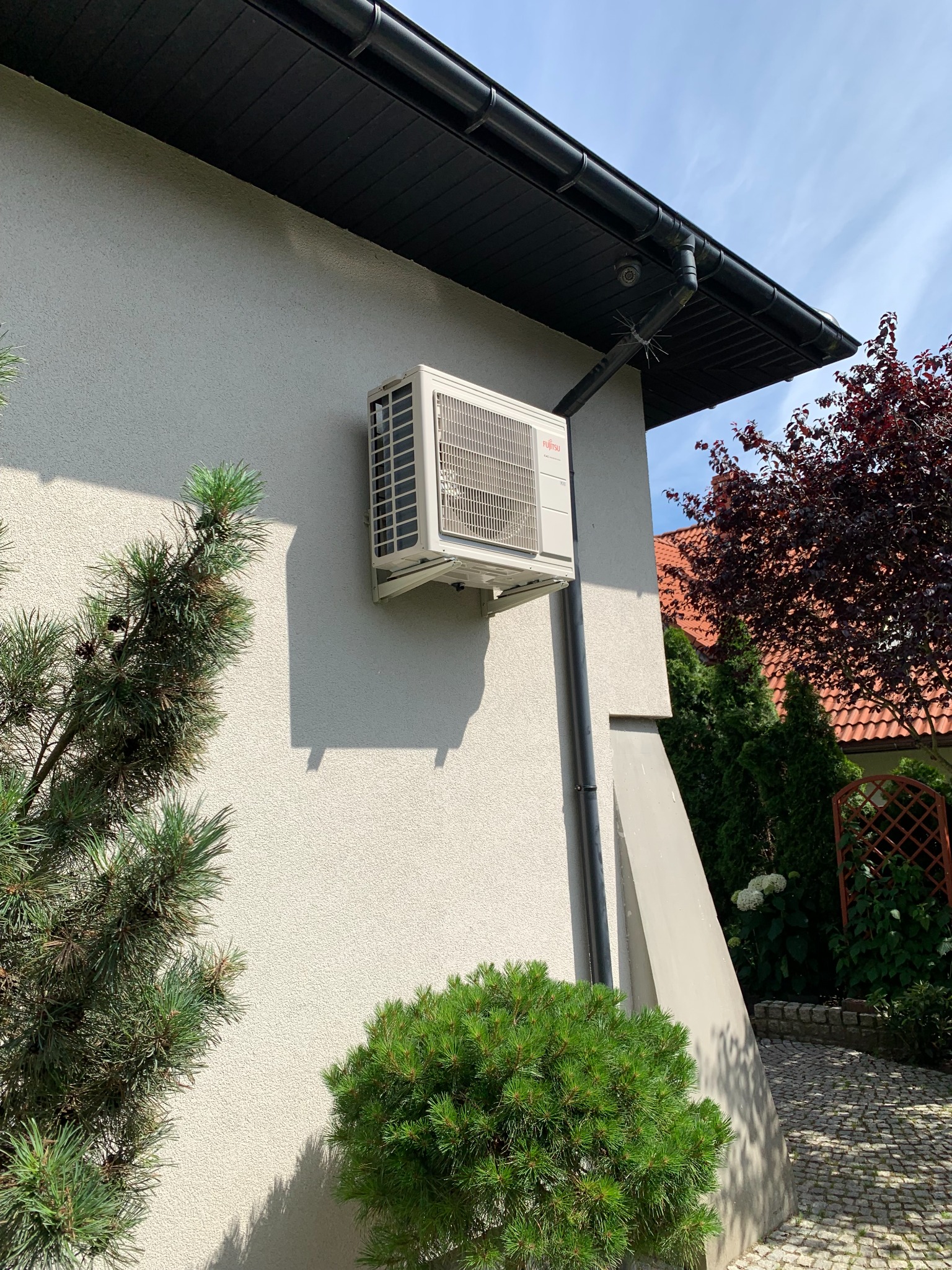 Zewnętrzna jednostka klimatyzacji Fujitsu zamontowana na jasnej ścianie domu, widoczna z boku, z elementami instalacji odprowadzającej wodę, na tle iglaków i czerwonego drzewa.