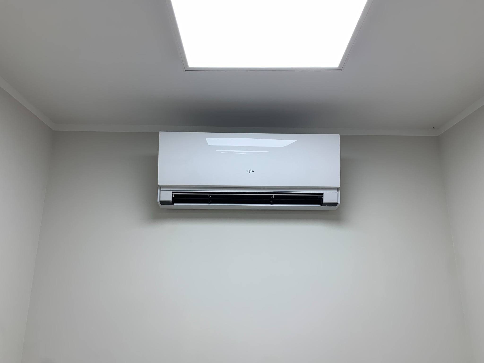 Nowoczesny, biały klimatyzator Fujitsu zamontowany na jasnoszarej ścianie pod sufitem w minimalistycznym wnętrzu biura lub domu, oświetlony przez kwadratową lampę sufitową.