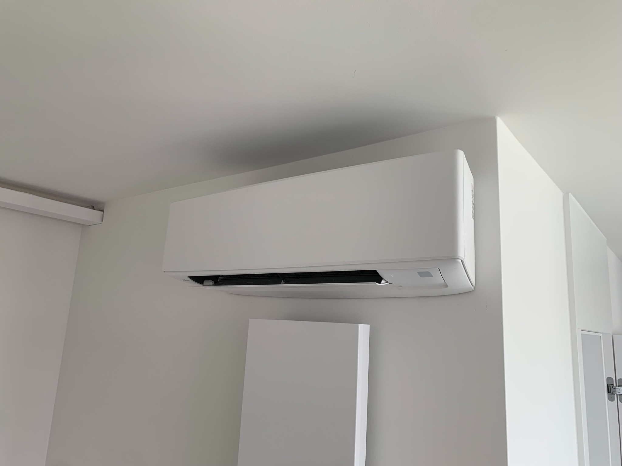 Biały klimatyzator ścienny zamontowany w minimalistycznym wnętrzu, widoczny fragment drzwi i panel dekoracyjny, oświetlenie sufitowe