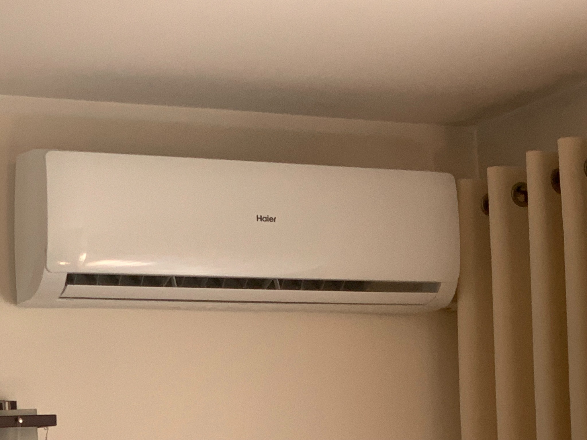 Świeżo zamontowany, biały klimatyzator Haier na ścianie w beżowym pomieszczeniu, widoczne zasłony w tle.