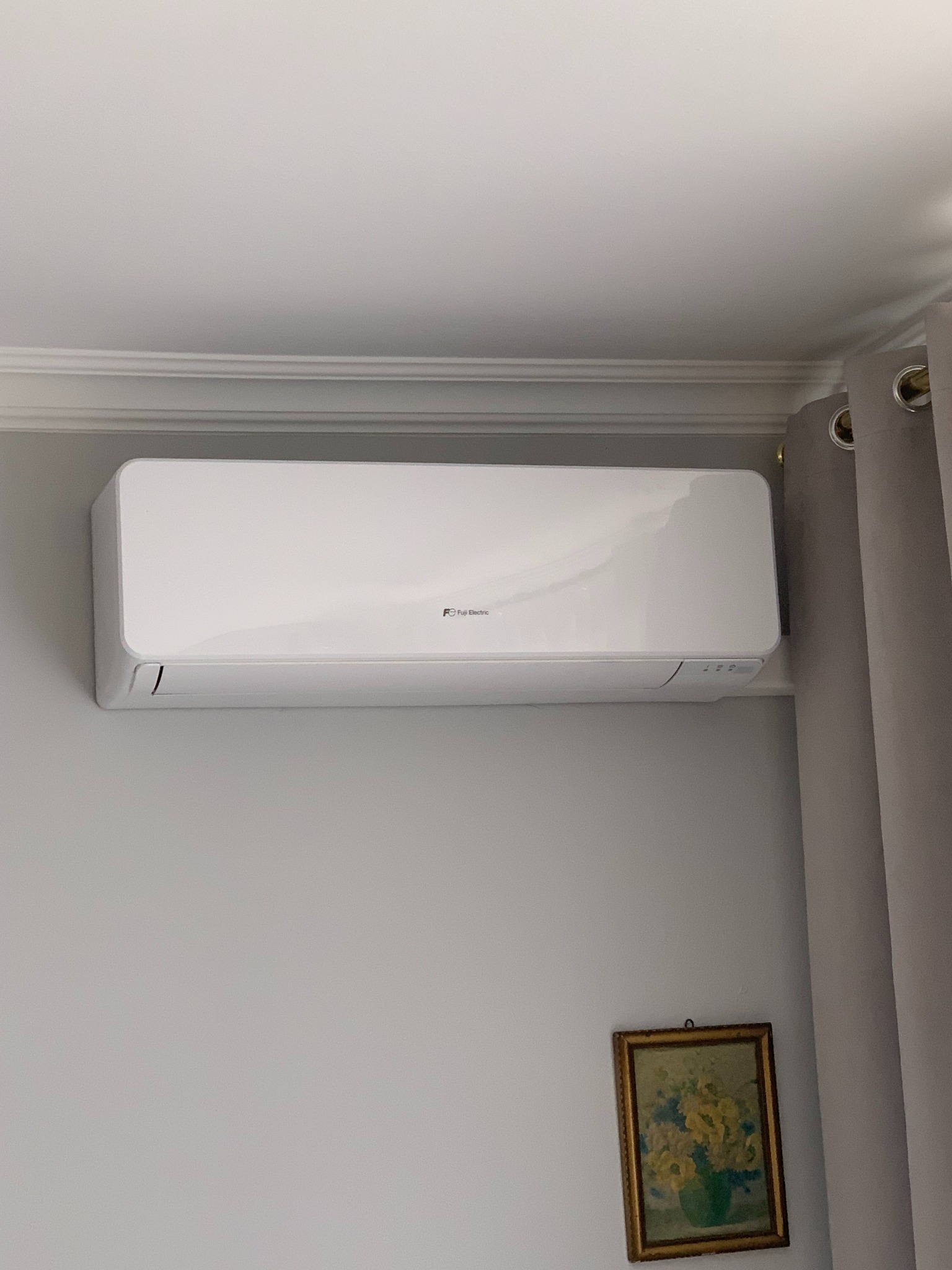 Biały klimatyzator Fuji Electric zamontowany na szarej ścianie w pomieszczeniu z listwą przysufitową i zasłoną, pod nim wisi obrazek w złotej ramie.