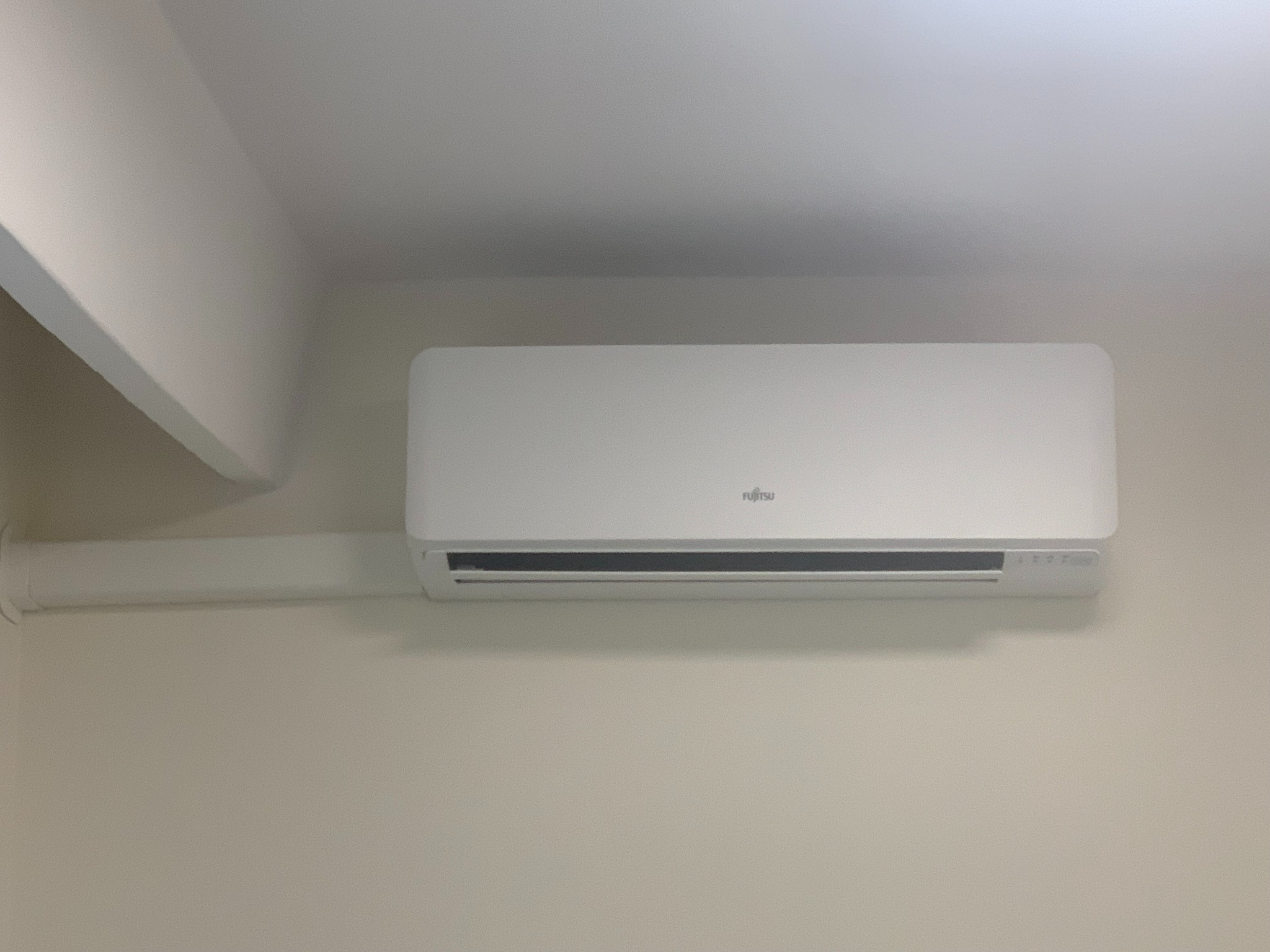 Biały klimatyzator Fujitsu zamontowany na jasnej ścianie w pomieszczeniu z widoczną rurą instalacyjną i fragmentem sufitu.