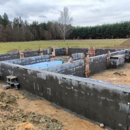 ZBYCHU-Usługi Wielobranżowe Zbigniew Słyż - Budowa fundamentów z bloczków betonowych na terenie niezabudowanym, widoczne zbrojenia, szalunki i pracownik w kombinezonie.