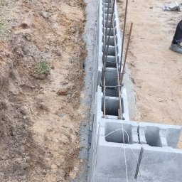 ZBYCHU-Usługi Wielobranżowe Zbigniew Słyż - Wykop pod fundamenty z częściowo wzniesioną ścianą z betonowych bloczków i wystającymi prętami zbrojeniowymi, widok z góry na plac budowy.