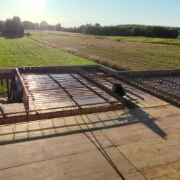 ZBYCHU-Usługi Wielobranżowe Zbigniew Słyż - Zbrojenie płyty fundamentowej na drewnianym szalunku, widok z góry na tle zielonego krajobrazu.