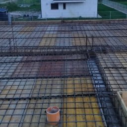 ZBYCHU-Usługi Wielobranżowe Zbigniew Słyż - Zbrojenie stropu budynku jednorodzinnego, widoczne pręty stalowe ułożone w kratę, szalunek z desek, pomarańczowe rury wentylacyjne wystające ponad konstrukcję, w tle dom w trakcie budowy.