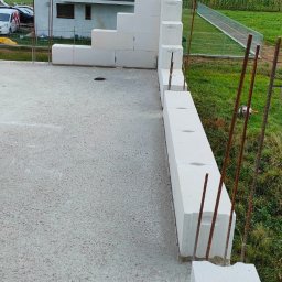 ZBYCHU-Usługi Wielobranżowe Zbigniew Słyż - Wznoszenie ściany z bloczków betonu komórkowego na fundamencie domu jednorodzinnego, widoczne pręty zbrojeniowe, w tle dom w stanie surowym otwartym i pole uprawne.
