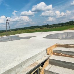 ZBYCHU-Usługi Wielobranżowe Zbigniew Słyż - Świeżo wylany betonowy fundament z widocznymi prętami zbrojeniowymi i kałużami wody, na tle błękitnego nieba z obłokami i polami uprawnymi.