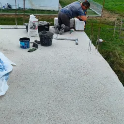 Pracownik na kolanach, używając poziomicy, układa bloczki na fundamencie domu w budowie, w tle widoczny dom w stanie surowym otwartym i zielone pole.