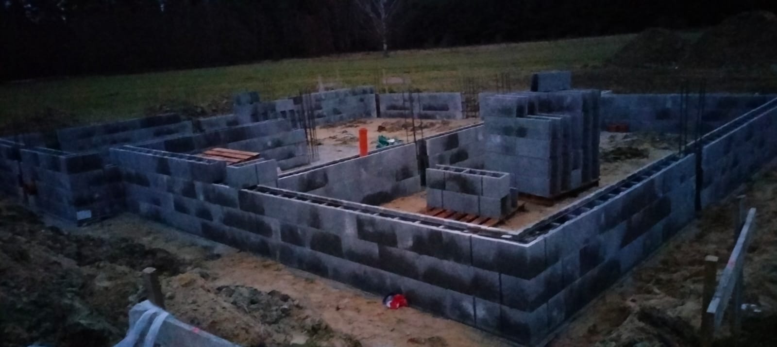 Wzniesione ściany fundamentowe z bloczków betonowych na działce budowlanej, widoczne wystające pręty zbrojeniowe i pomarańczowa rura.