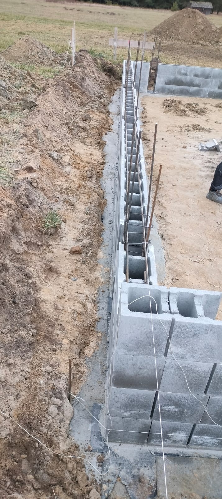 Wykop pod fundamenty z częściowo wzniesioną ścianą z betonowych bloczków i wystającymi prętami zbrojeniowymi, widok z góry na plac budowy.