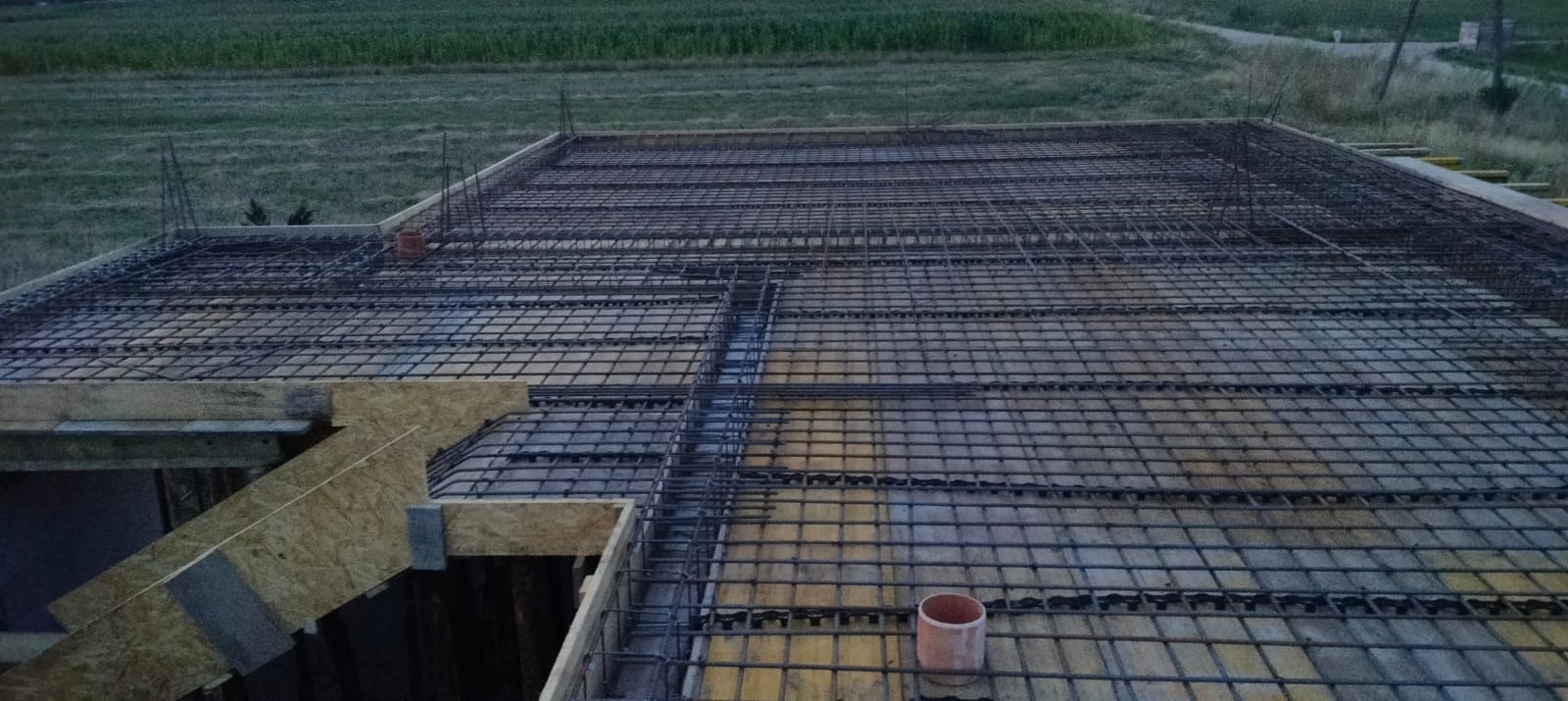 Zbrojenie płyty fundamentowej z widoczną konstrukcją szalunkową i wystającymi prętami zbrojeniowymi w tle zielone pole.