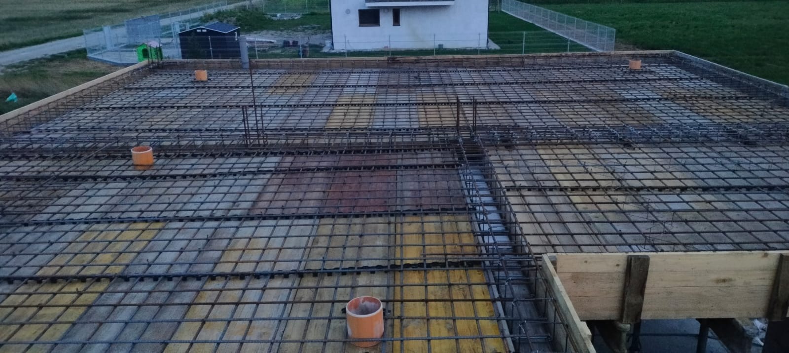 Zbrojenie stropu budynku jednorodzinnego, widoczne pręty stalowe ułożone w kratę, szalunek z desek, pomarańczowe rury wentylacyjne wystające ponad konstrukcję, w tle dom w trakcie budowy.