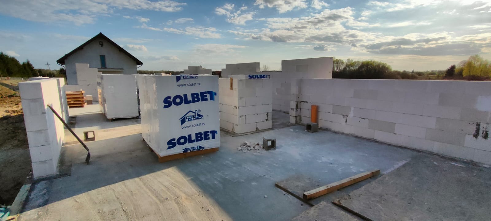 Wznoszone ściany domu z betonu komórkowego Solbet, widoczne palety z materiałem, całość na fundamencie, w tle dom i drzewa.
