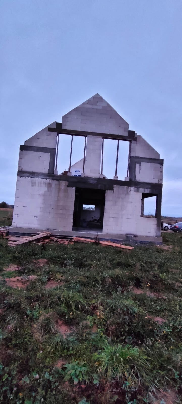 Surowy stan domu jednorodzinnego z bloczków, widoczne otwory okienne i drzwiowe, teren zarośnięty trawą, pochmurne niebo w tle.