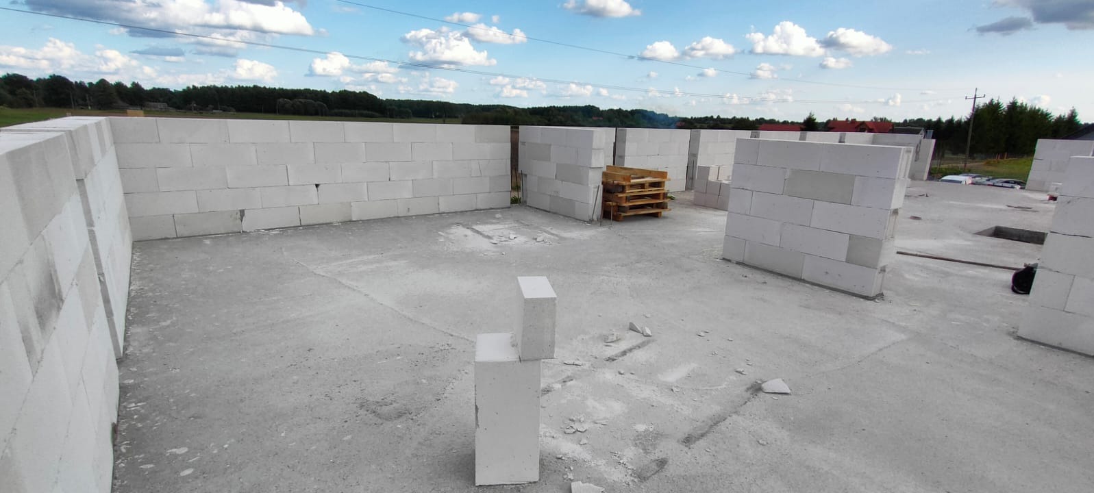 Wznoszone ściany z białych bloczków na fundamencie domu jednorodzinnego, widoczne palety i zarys terenu w tle.