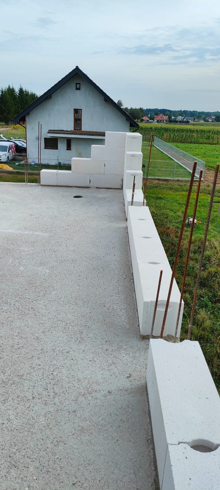 Wznoszenie ściany z bloczków betonu komórkowego na fundamencie domu jednorodzinnego, widoczne pręty zbrojeniowe, w tle dom w stanie surowym otwartym i pole uprawne.