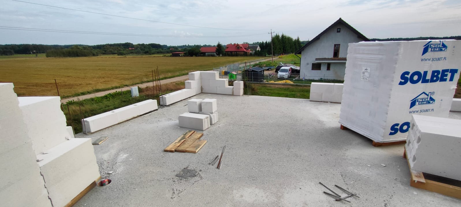 Wznoszenie ścian z białych bloczków Solbet na fundamencie domu jednorodzinnego, widok na okolicę z polami i zabudowaniami.