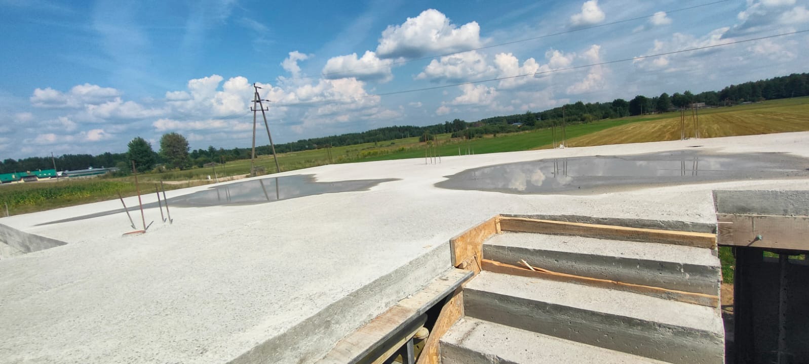 Świeżo wylany betonowy fundament z widocznymi prętami zbrojeniowymi i kałużami wody, na tle błękitnego nieba z obłokami i polami uprawnymi.