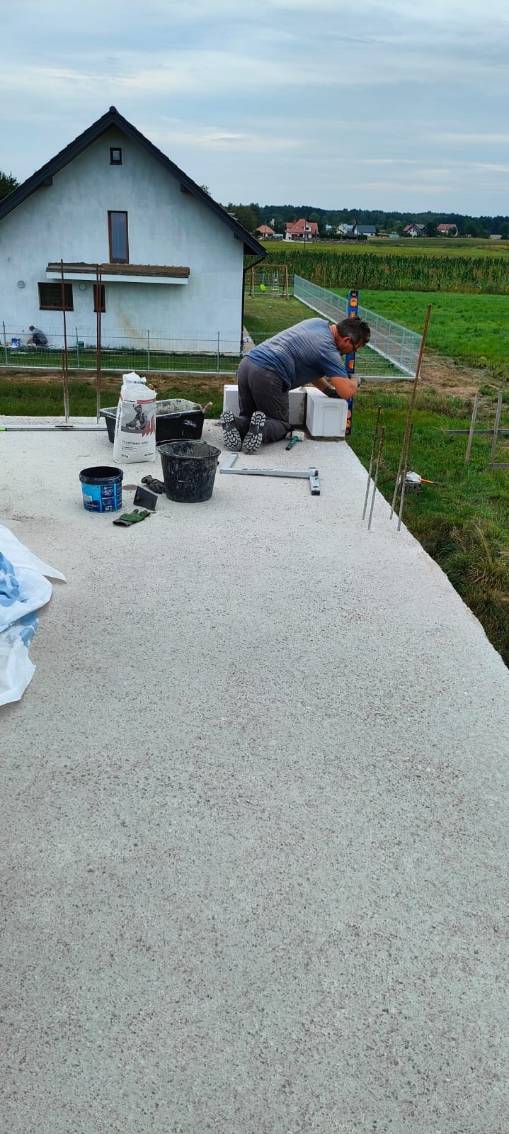 Pracownik na kolanach, używając poziomicy, układa bloczki na fundamencie domu w budowie, w tle widoczny dom w stanie surowym otwartym i zielone pole.