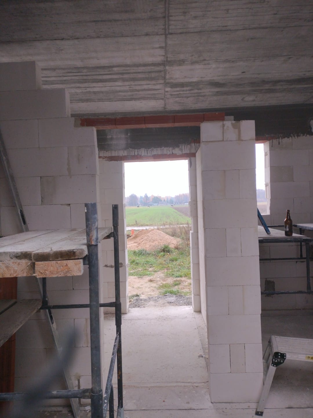 Wnętrze surowego domu w trakcie budowy, ściany wzniesione z białych bloczków, widoczne rusztowania i butelka na parapecie, w tle krajobraz polny.