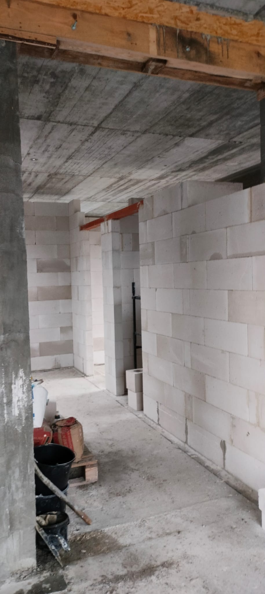 Wnętrze budynku w trakcie wznoszenia ścian z białych bloczków, widoczne surowe betonowe stropy i prowizoryczne drewniane wzmocnienia, na podłodze narzędzia i materiały budowlane.