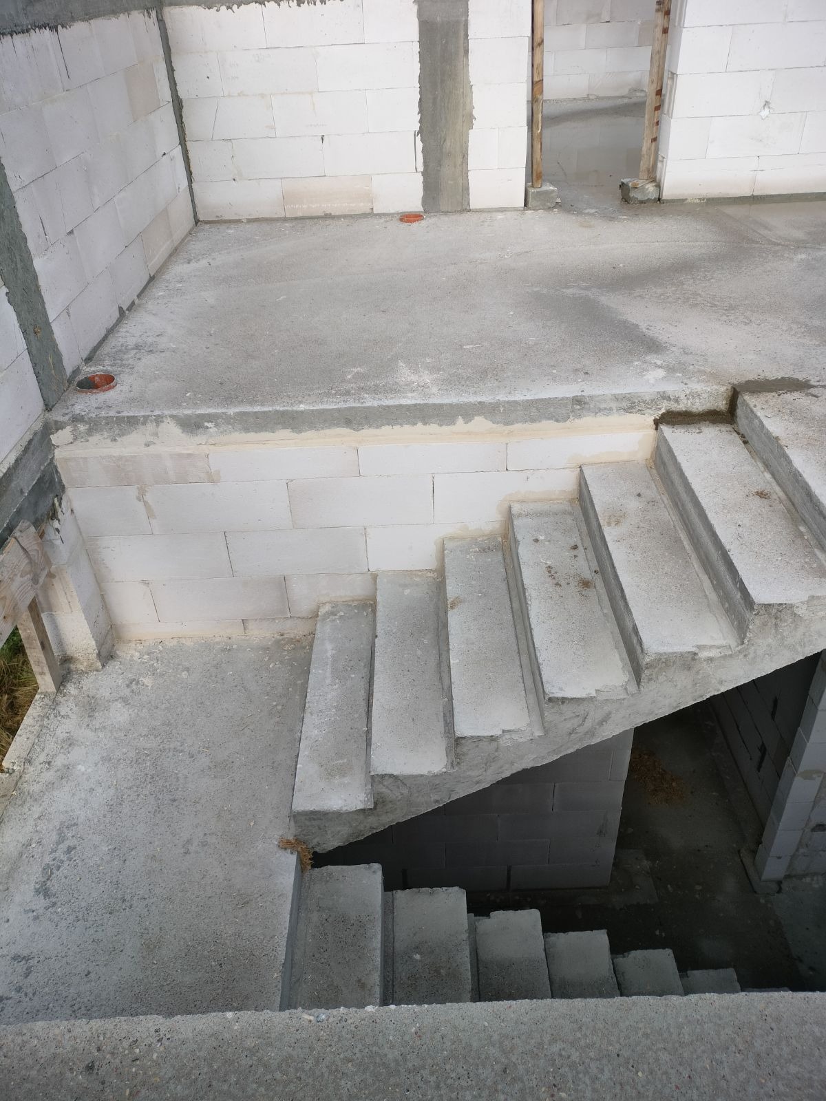 Surowe schody betonowe wewnątrz budynku w stanie surowym, widoczne ściany z białego betonu komórkowego i surowy betonowy strop, fragment instalacji kanalizacyjnej w podłodze.
