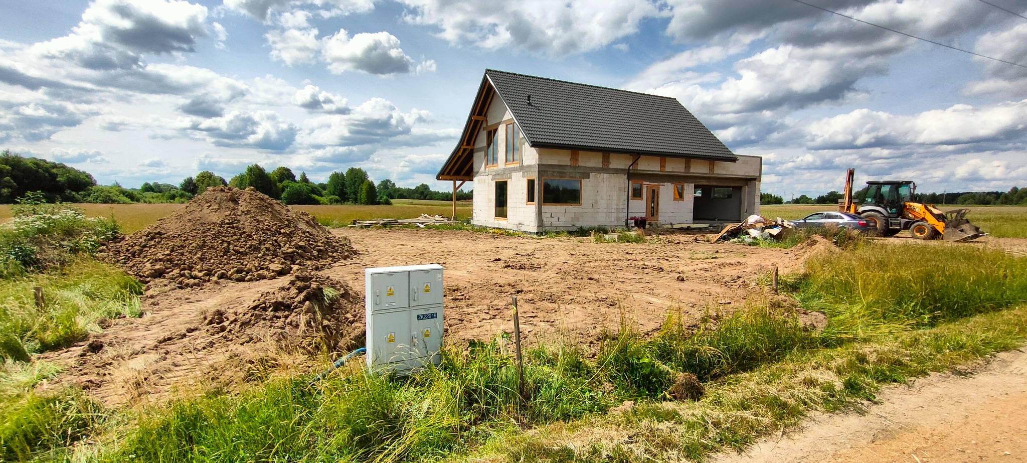 Surowy stan domu jednorodzinnego z bloczków, z czarną dachówką, widoczne okna i drzwi bez wykończenia, na pierwszym planie skrzynka elektryczna, w tle koparko-ładowarka i samochód